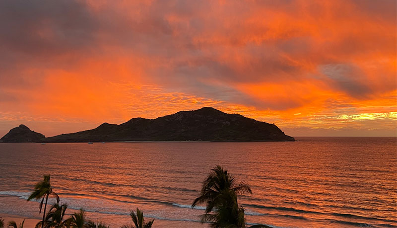 A coastal sunset view in Mazatlán, Mexico, featuring a dark mountain silhouette in the middle of a calm ocean, with a vibrant orange and cloudy sky reflected on the water and palm trees in the foreground.