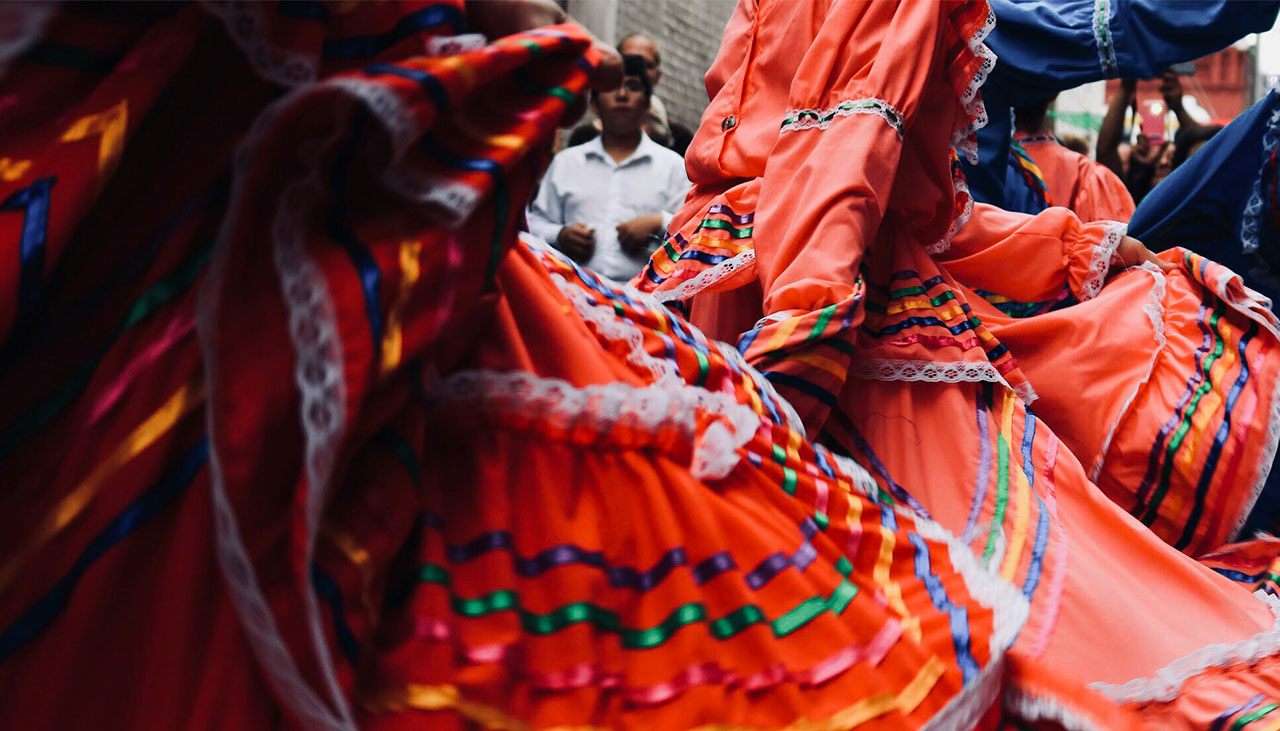 A vibrant scene of Mexican folk dancers in traditional orange and red Jalisco-style dresses, performing in a historic plaza with a group of musicians in the background.