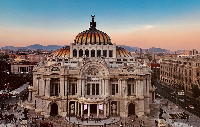 The Palacio de Bellas Artes in Mexico City at sunset, featuring its iconic orange and yellow domed roof and ornate Neoclassical white marble exterior.