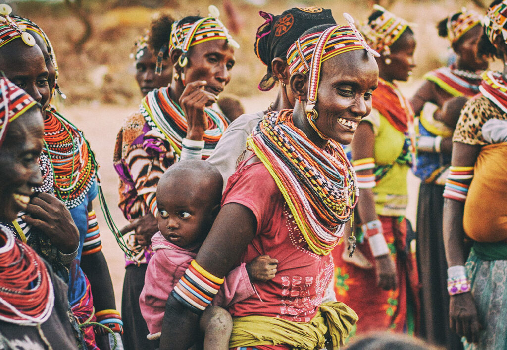 A group of Samburu people in Kenya, including a woman with elaborate beaded necklaces and a young child, gathered together during a traditional community ceremony in a rural landscape.
