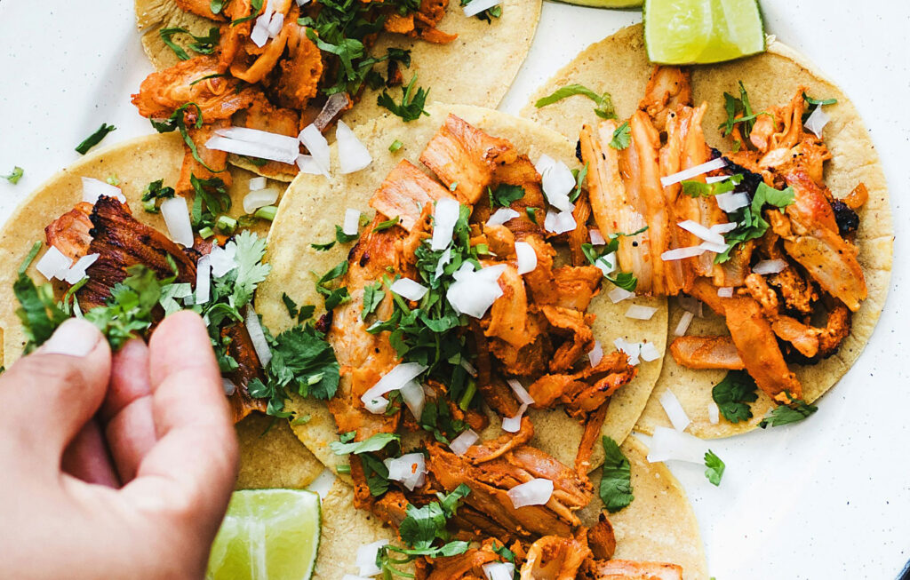 A close-up of two traditional Mexican street tacos on a plate, topped with fresh cilantro, onions, and white cheese, with a hand squeezing a fresh lime wedge over the seasoned meat.