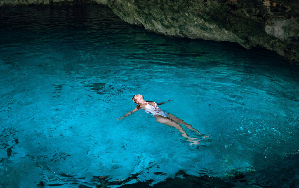 Alt Text: A woman swimming in the clear, turquoise water of a sunlit Mexican cenote, surrounded by limestone cave walls and lush greenery.