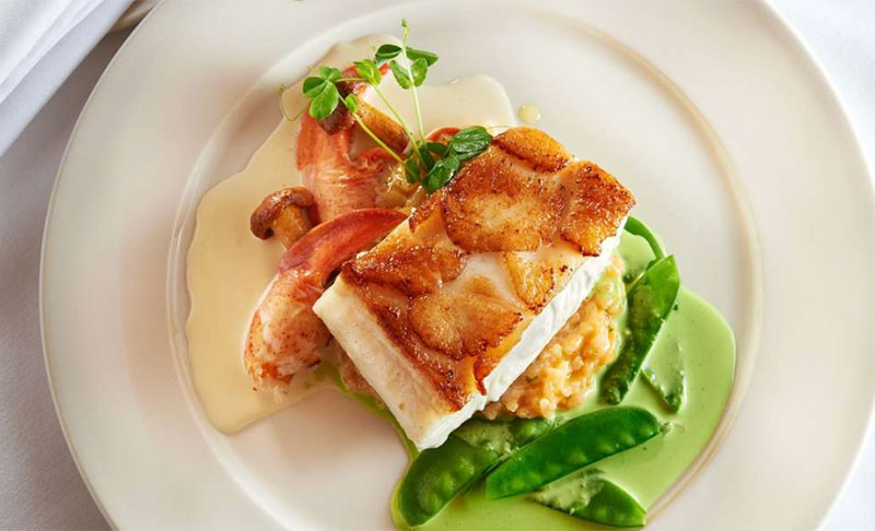 A gourmet seafood dish from GW Fins featuring their signature "Scalibut"—halibut wrapped in thin slices of sea scallop, served with a vibrant vegetable medley and a delicate savory sauce.