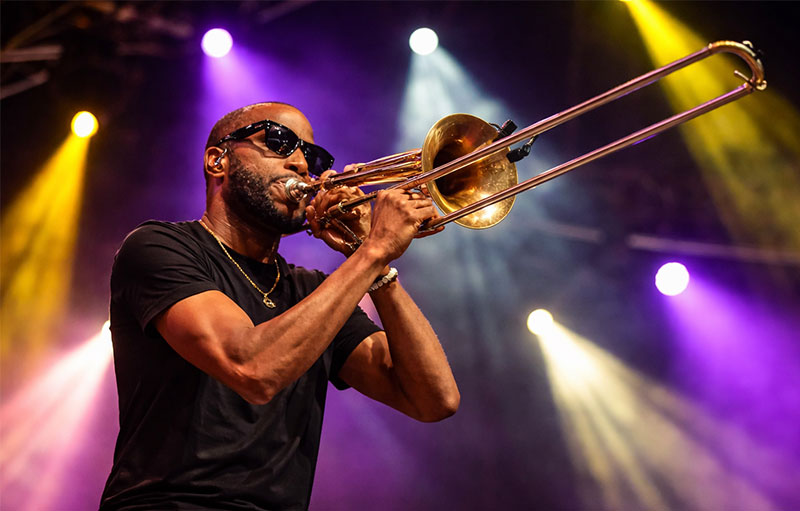 Trombone Shorty performing live at New Orleans Jazz Fest with colorful stage lights and energetic crowd atmosphere