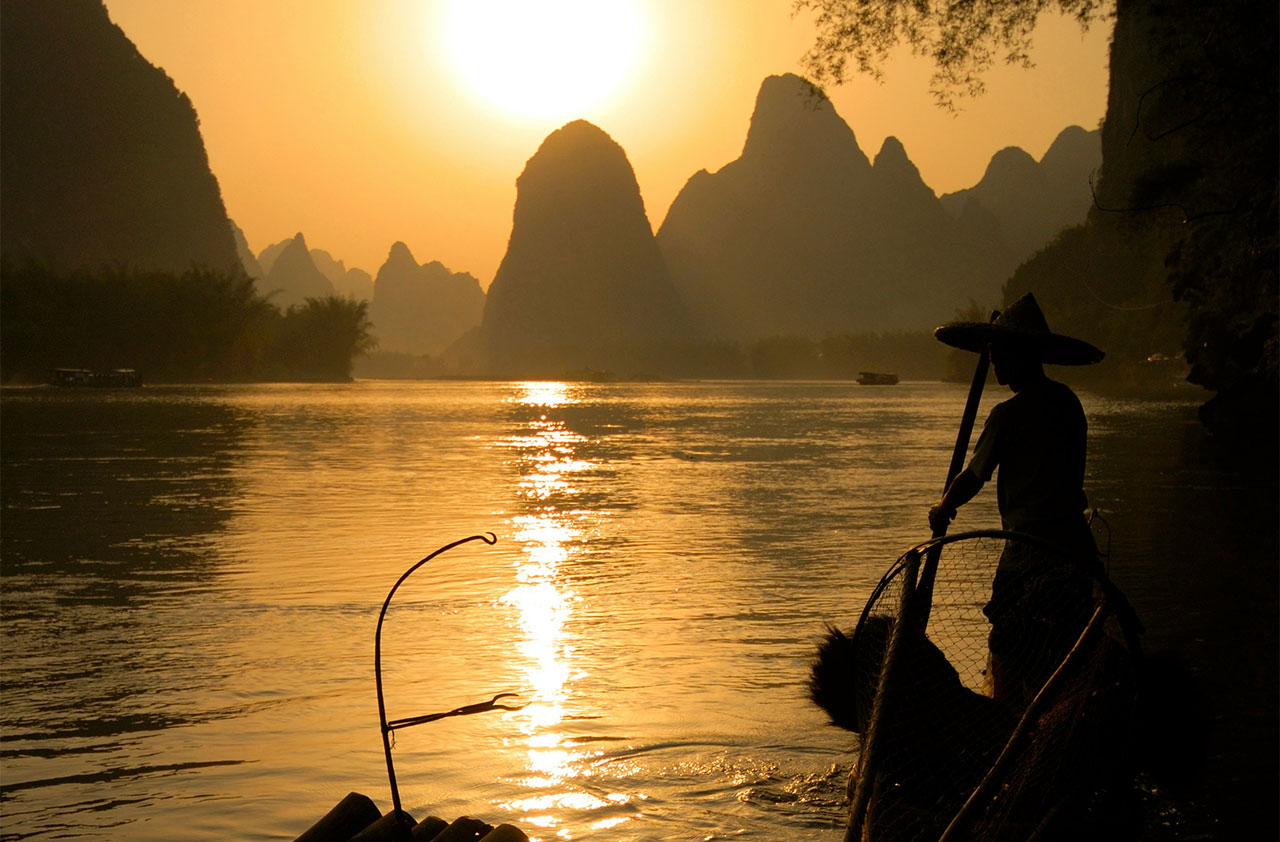 A silhouette of a fisherman in a traditional conical hat standing on a wooden boat, holding a large net over a calm river at sunset, with karst mountains in the background.