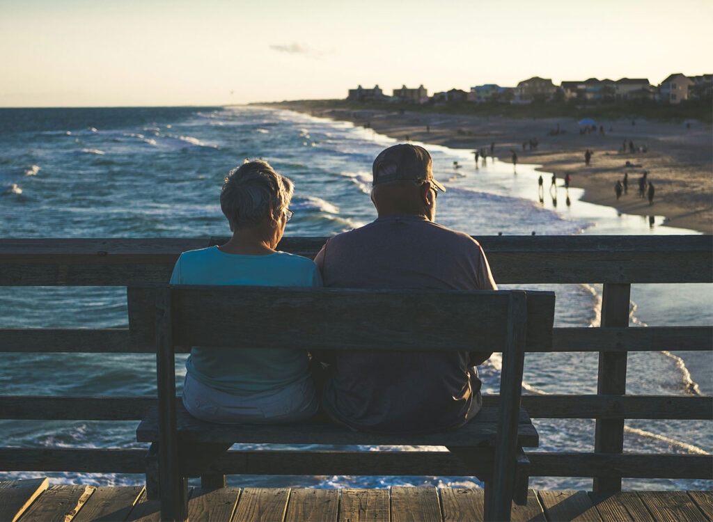 A retired couple sitting on a wooden bench on a pier, looking out over a calm ocean at sunset.