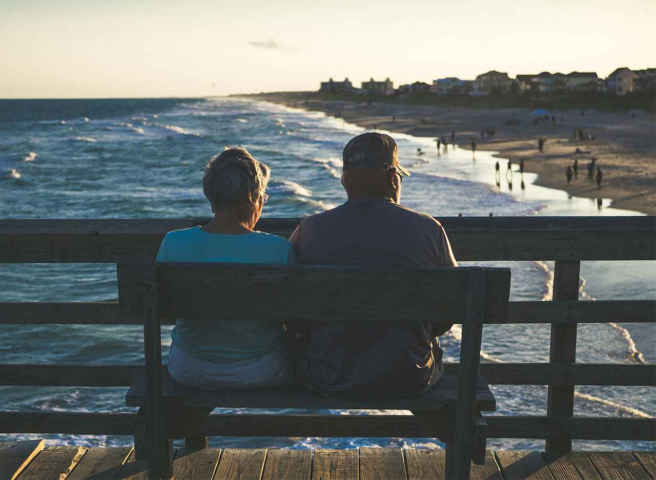 A retired couple sitting on a wooden bench on a pier, looking out over a calm ocean at sunset.