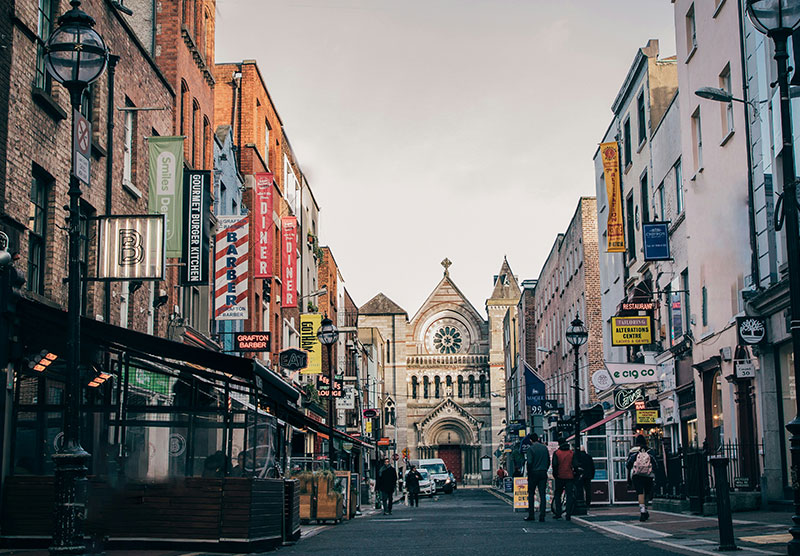A vibrant street scene on Anne Street in Dublin, showing the pedestrianized lane lined with colorful flower baskets, historic brick buildings, and the prominent blue and white storefront of Sheridan’s Cheesemongers