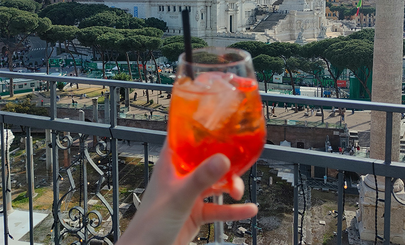 A vibrant, close-up shot focuses on a bright orange Aperol Spritz in a large wine glass, held up against a Roman backdrop.