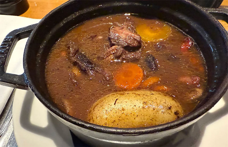 A classic and comforting presentation of Beef Bourguignon at Au Bourguignon du Marais, featuring tender chunks of slow-cooked beef in a rich, dark wine sauce, garnished with pearl onions, mushrooms, and vibrant green peas, served alongside a side of golden mashed potatoes.