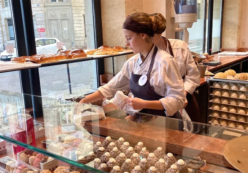 A view through a bakery window of two pastry chefs in white uniforms and aprons meticulously assembling "Le Merveilleux" cakes, with rows of small meringue bases visible on the wooden counter in the foreground.