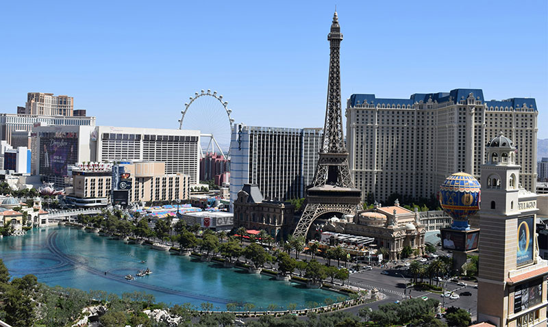 high-angle view from a balcony at the Cosmopolitan looking over the Las Vegas Strip, featuring the Eiffel Tower at Paris Las Vegas, the High Roller observation wheel, and the surrounding resort architecture under a clear blue sky.