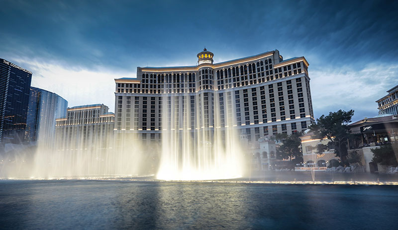 A wide shot of the Bellagio Hotel and Casino at dusk, with the fountains in the foreground shooting high plumes of water into the air against a deep blue twilight sky.