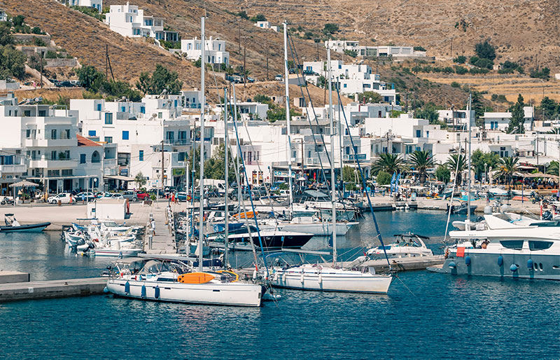 A wide-angle shot of a harbor in a Greek island village, featuring several traditional white and blue fishing boats moored in turquoise water, with whitewashed buildings and a sun-drenched coastal hillside in the background.