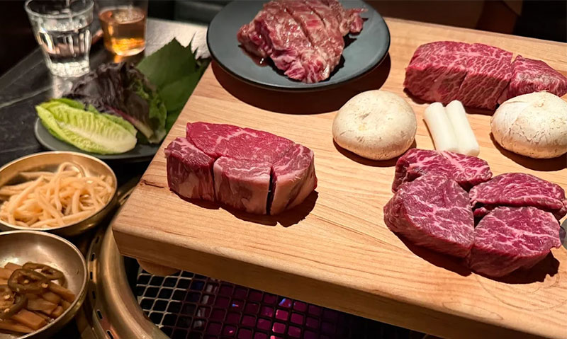A sophisticated, dimly lit interior of COTE Korean Steakhouse in Las Vegas, featuring round dining tables with built-in grills under dramatic, modern overhead lighting.