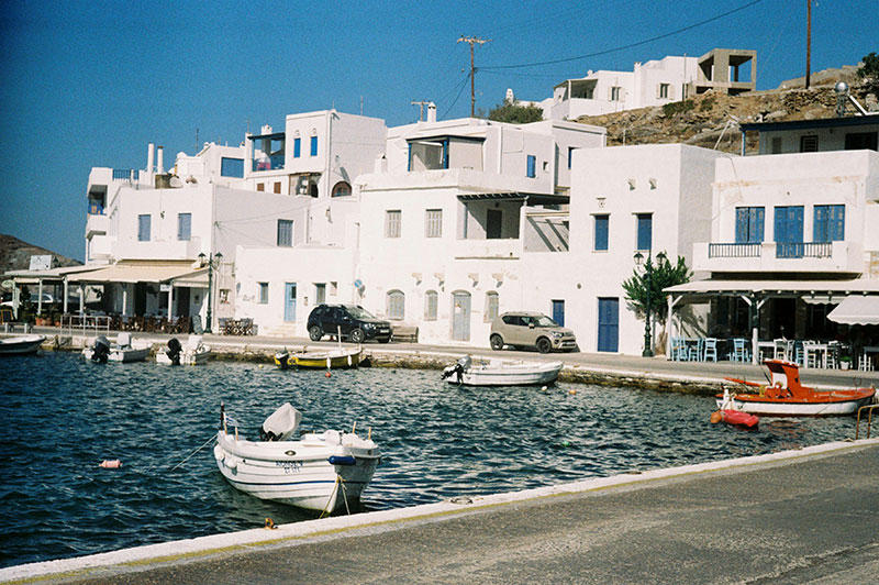 A peaceful view of a traditional Greek island port in Tinos, featuring a cluster of white-washed buildings with blue windows, a small fishing boat moored in the calm turquoise water, and a quiet waterfront walkway.