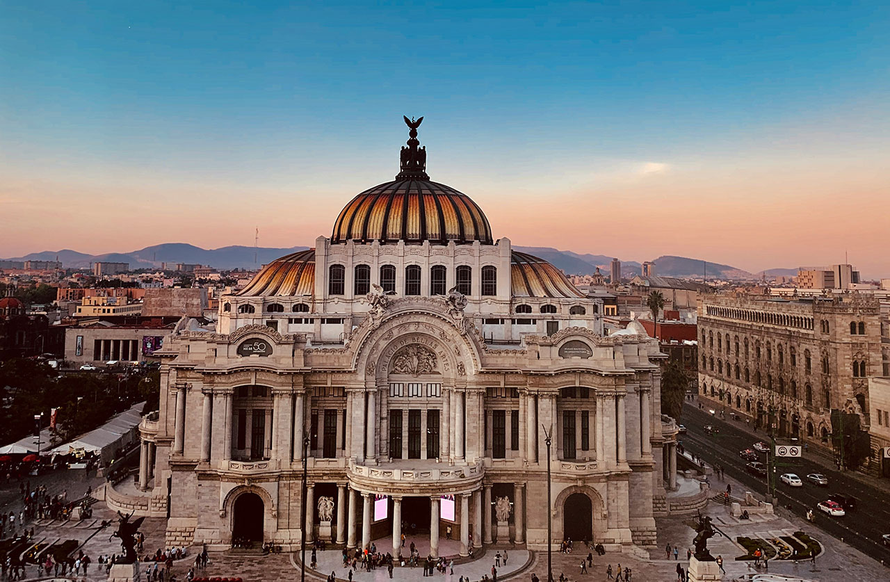 A wide-angle sunset view of the Palacio de Bellas Artes in Mexico City, featuring its iconic orange and yellow domed roof against a soft pink and blue sky