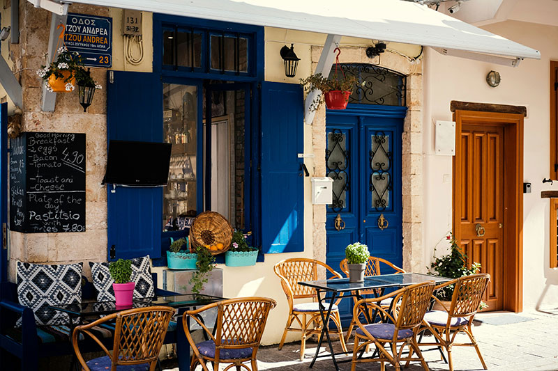 A high-angle, scenic view of the Chania Venetian Harbor on the island of Crete, featuring the iconic 16th-century lighthouse, colorful waterfront buildings, and traditional wooden boats moored in the calm, turquoise water.