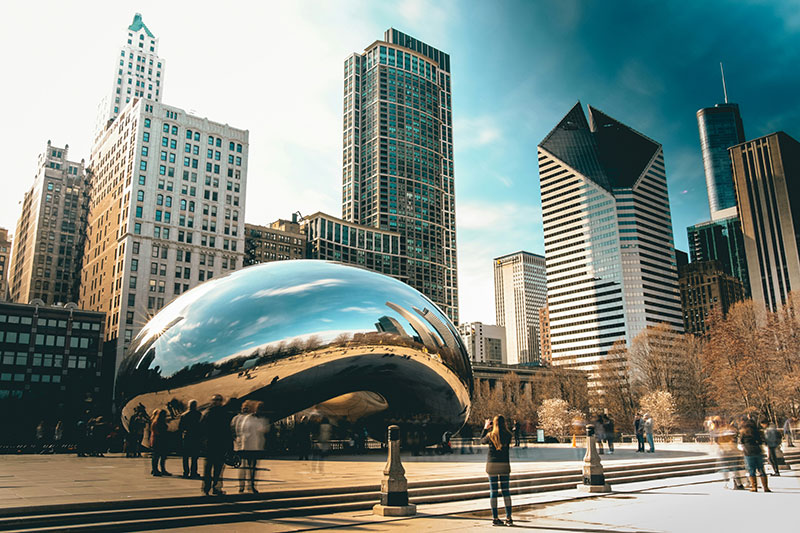 A view of the Chicago skyline and high-rise buildings reflecting in the polished, mirror-like surface of the Cloud Gate sculpture (the "Bean") in Millennium Park under a clear blue sky.