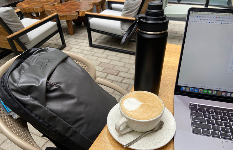 A close-up, overhead shot of a wooden table featuring a white cup of cappuccino with latte art, a black thermal water bottle, and an open laptop, representing a typical remote work setup for a digital nomad in Colombia.