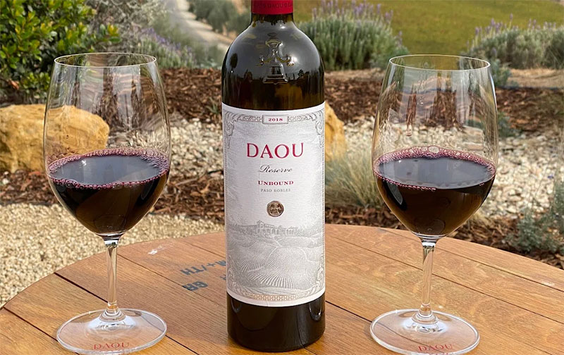 A bottle of DAOU Cabernet Sauvignon and two glasses of red wine on an outdoor table overlooking the rolling vineyard hills of Paso Robles.
