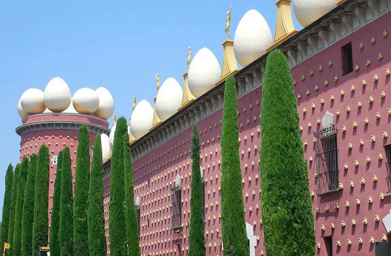The exterior of the Dalí Theatre-Museum in Figueres, Spain, featuring a red facade adorned with small golden bread sculptures and large white eggs lining the rooftop.