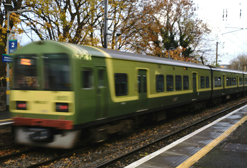 A long exposure shot of a green and white DART train blurred in motion as it pulls away from a station platform in Dublin, with the historic stone station walls and modern yellow platform markings visible