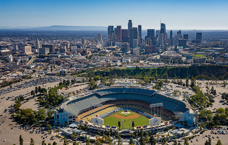 A wide-angle sunset view of Dodger Stadium in Los Angeles, showing the bright green field and blue stadium seating with the downtown city skyline and mountains visible in the hazy distance.