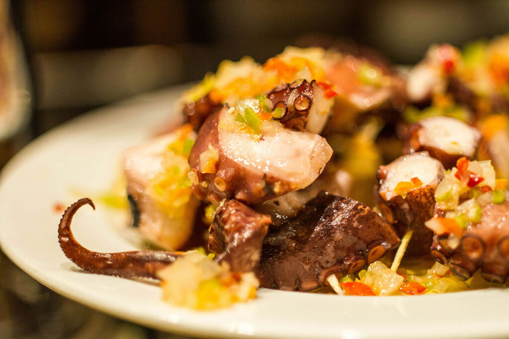 A close-up shot of a traditional Basque pintxo featuring tender pieces of grilled octopus seasoned with paprika, served over a slice of potato on a crusty piece of bread.