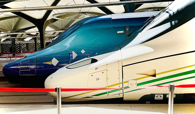 A sleek white and green Talgo 350 high-speed train, known as the "Haramain Express" or "AVE of the Desert," featuring its distinctive aerodynamic "duck-nose" front, traveling through the desert landscape between Mecca and Medina.