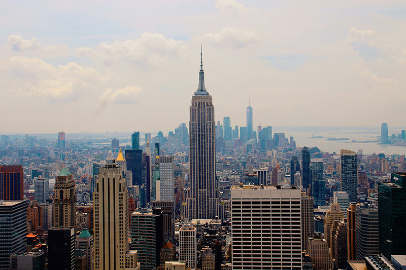 A high-angle aerial view of the New York City skyline during the day, with the iconic Empire State Building rising prominently above a dense grid of skyscrapers stretching toward the hazy horizon.