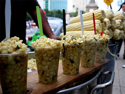 A close-up of several cups of esquites (Mexican street corn) topped with cream, cheese, and chili powder, sitting on a vendor's stall in Mexico City.