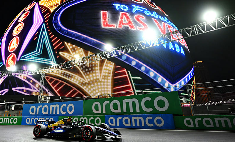 An Formula 1 race car speeding past the "Welcome to Fabulous Las Vegas" sign at night, with motion-blurred neon lights and race branding in the background.