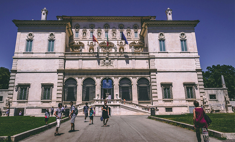 Galleria Borghese’s grand facade in Rome. The seventeenth-century villa features a symmetrical, light-ochre exterior adorned with classical sculptures in recessed niches and intricate stone relief carvings