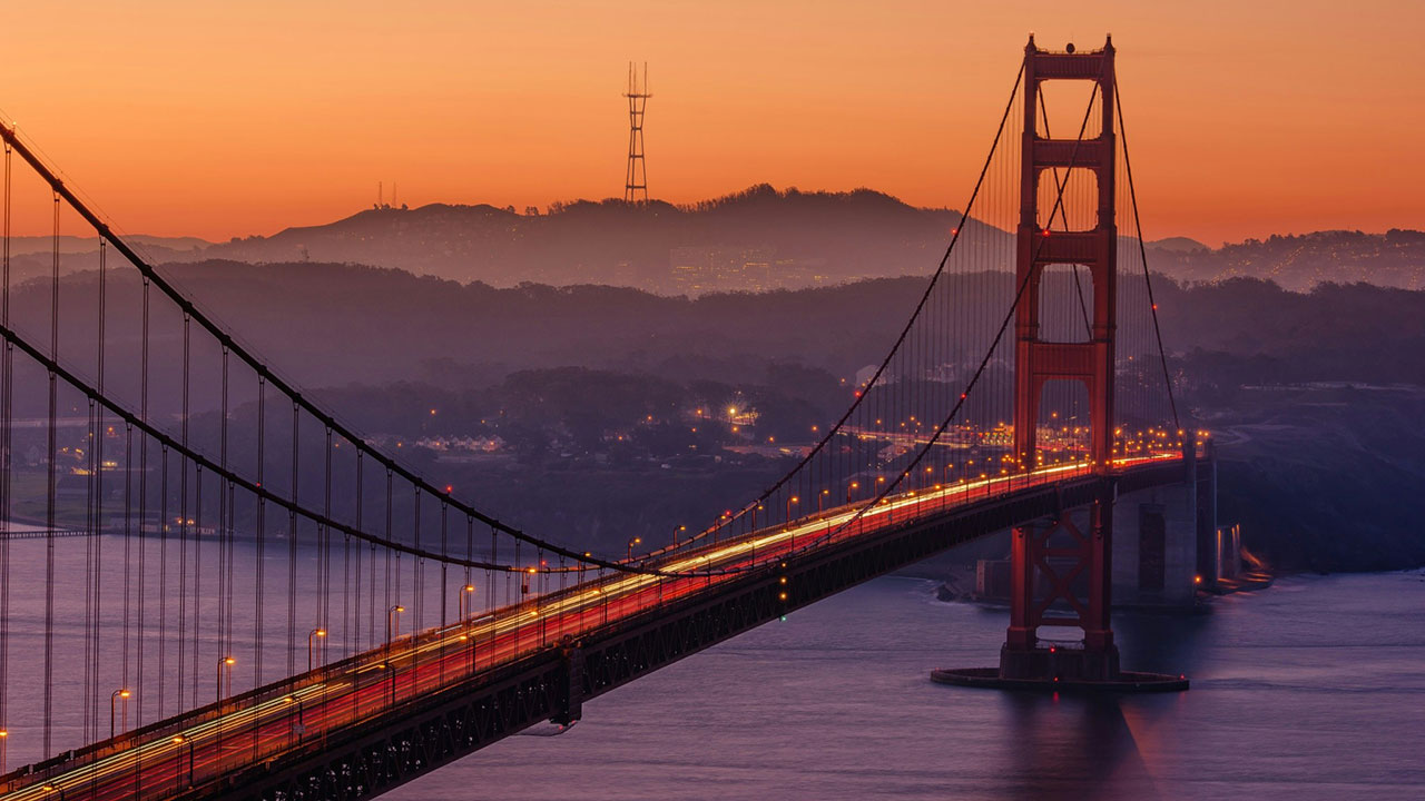 A wide-angle, scenic view of the Golden Gate Bridge at sunset, with the iconic orange towers glowing against a soft pink and purple sky and the calm waters of the bay in the foreground.