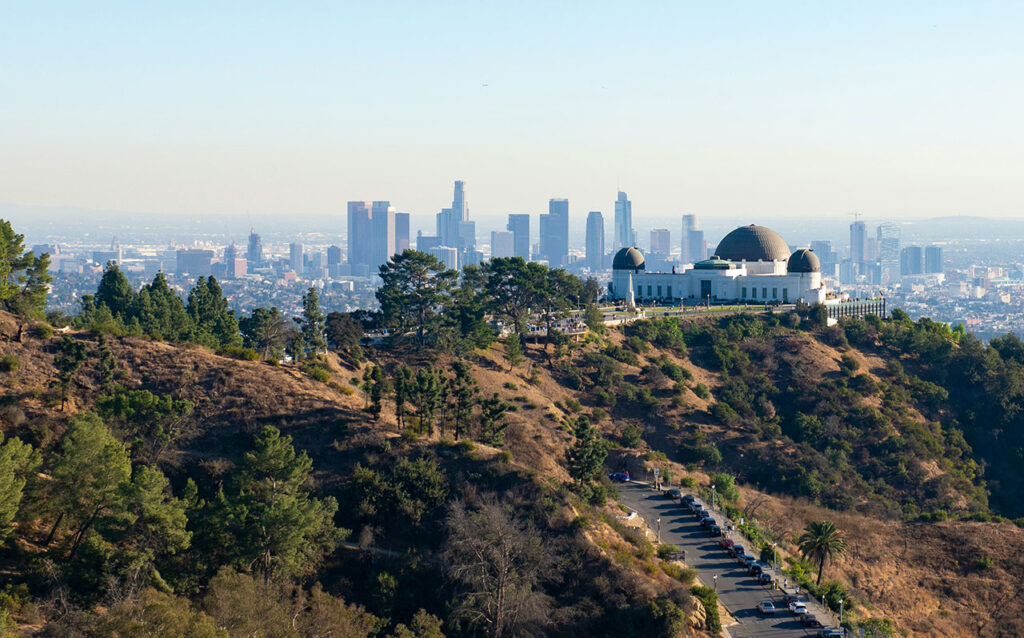 A panoramic view of the Los Angeles skyline at dusk, showing the dense city grid stretching toward the horizon with the silhouettes of the San Gabriel Mountains in the distance under a hazy sky.