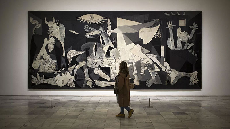 A wide-angle black and white photograph of visitors standing in front of Pablo Picasso's massive mural, Guernica, at the Reina Sofía Museum in Madrid.