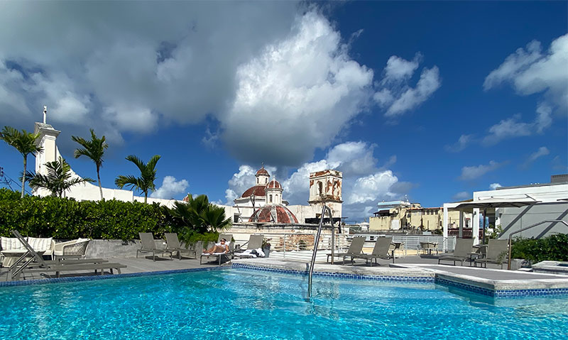 A sunny rooftop pool deck with lounge chairs overlooking the historic Cathedral of San Juan Bautista in Old San Juan.