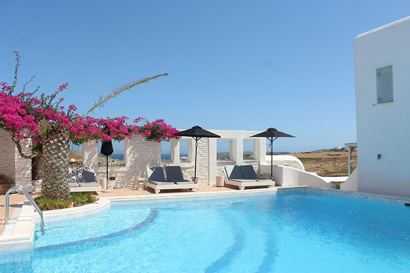A bright, sun-drenched outdoor patio of a boutique hotel in Paros, featuring a sparkling blue swimming pool surrounded by white stone walls, comfortable lounge chairs, and vibrant pink bougainvillea.