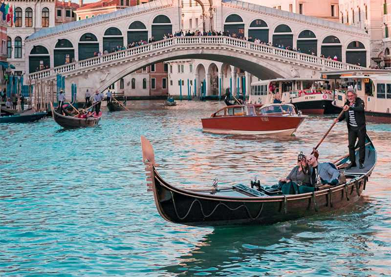 A couple enjoying a private gondola ride along a scenic Venetian canal, passing under a historic stone bridge at sunset.