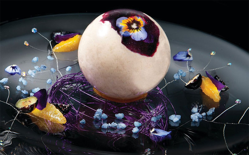 A close-up of a gourmet dish from Joël Robuchon, featuring a single, beautifully plated scallop topped with caviar and delicate garnishes on a dark, textured plate.