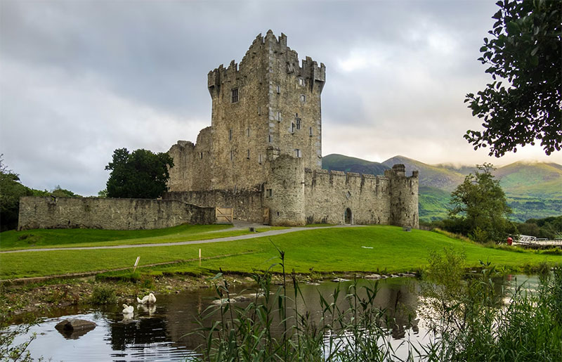 A striking view of Ross Castle in Killarney, featuring the medieval stone tower house standing tall against a backdrop of the serene Lough Leane and the distant, mist-covered mountains of Killarney National Park.