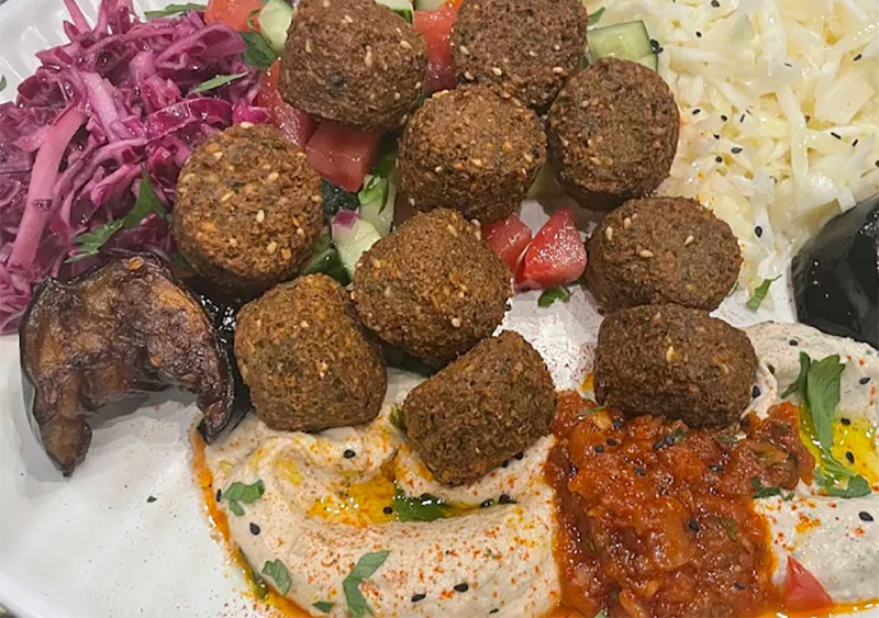 A close-up, appetizing shot of a signature falafel pita from L'As du Fallafel, overflowing with golden-brown falafel balls, bright purple cabbage, fresh cucumbers, and a generous drizzle of creamy tahini sauce.