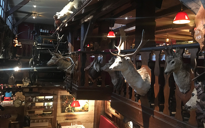 The rustic wooden interior of a traditional Savoyard restaurant in Chamonix, heavily decorated with taxidermy animals mounted on the walls and balcony railings. Caption: A traditional Savoyard tavern is the perfect setting for a classic mountain comfort meal. Expect eccentric, cozy interiors and an incredible amount of melted cheese.