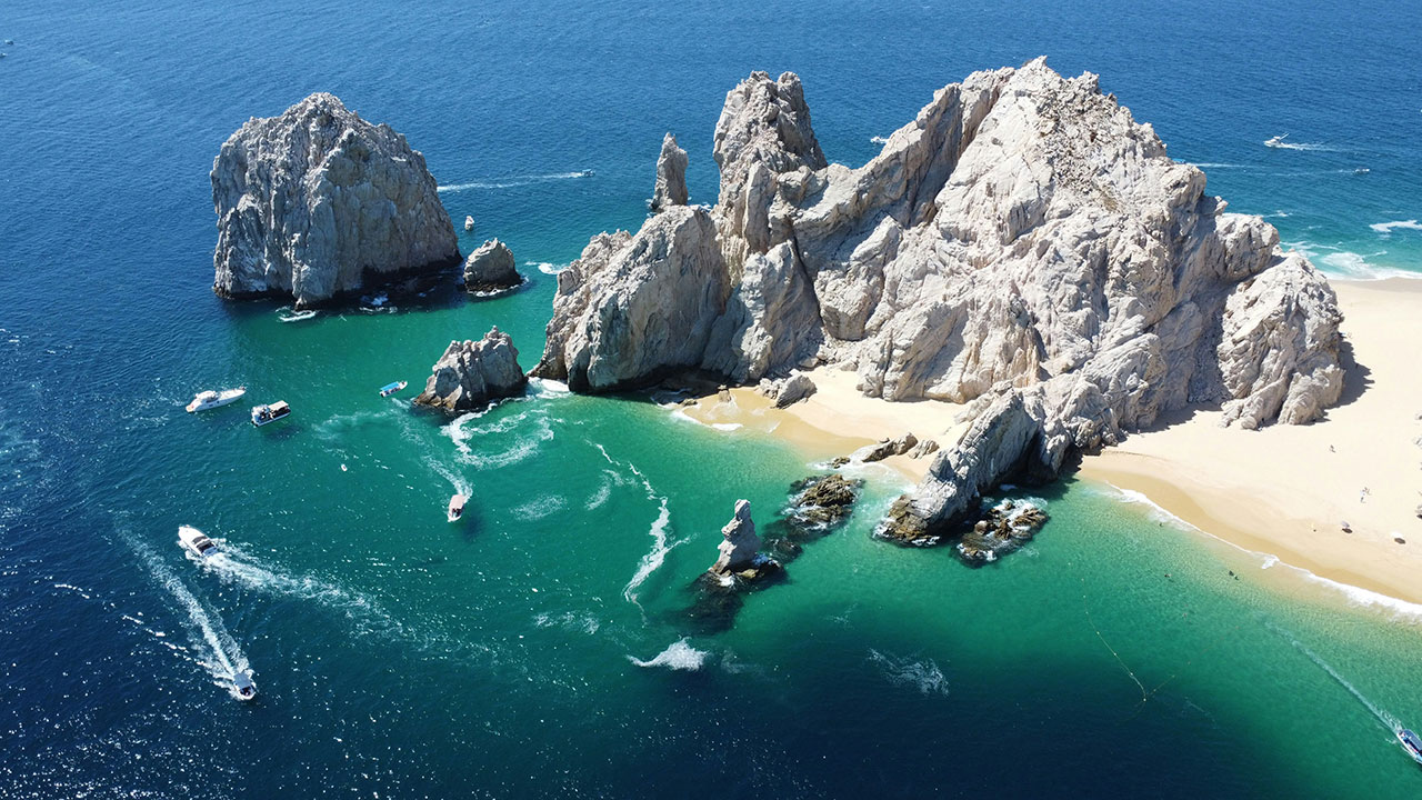 An aerial view of the iconic rugged rock formations at Land's End in Cabo San Lucas, surrounded by clear turquoise water and a hidden sandy beach.