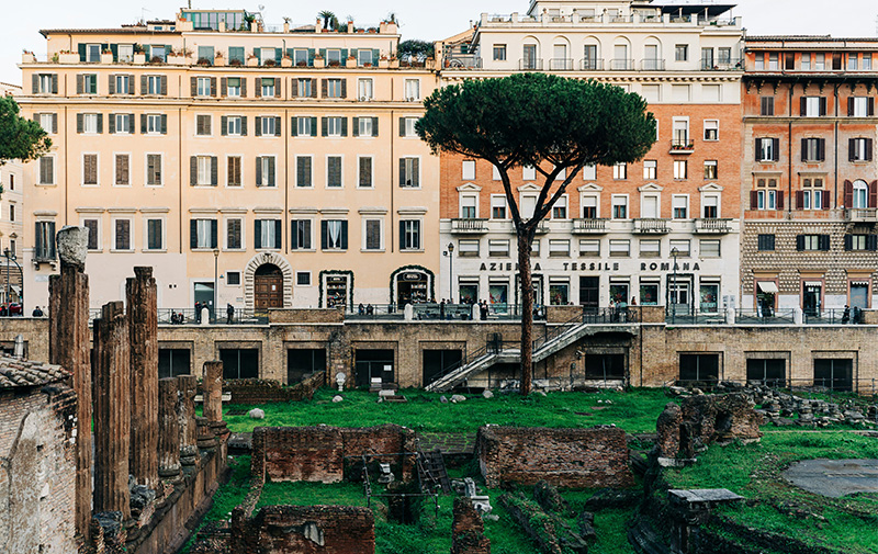 A medium-wide, slightly elevated shot of the Largo di Torre Argentina archaeological site in Rome. The rectangular sunken area features the ancient stone remains of four Republican-era temples, including weathered tuff and travertine columns, broken pedestals, and brick foundations