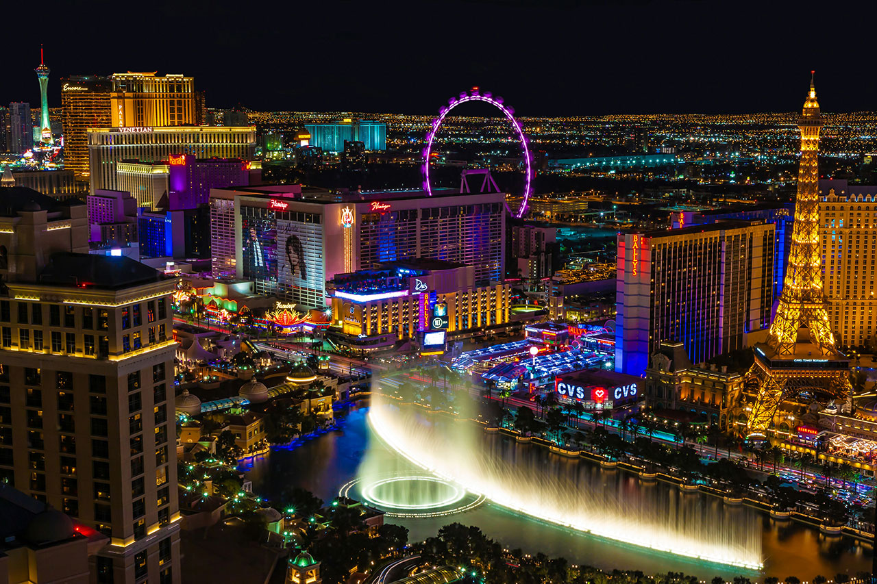 A high-angle nighttime view of the Las Vegas Strip featuring the High Roller observation wheel, the Eiffel Tower at Paris Las Vegas, and the glowing neon lights of various resort hotels and casinos.