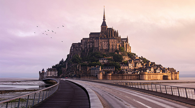 Wide shot of the historic island commune of Le Mont-Saint-Michel in Normandy, France, featuring the medieval abbey rising above the tidal flats under a soft sky.