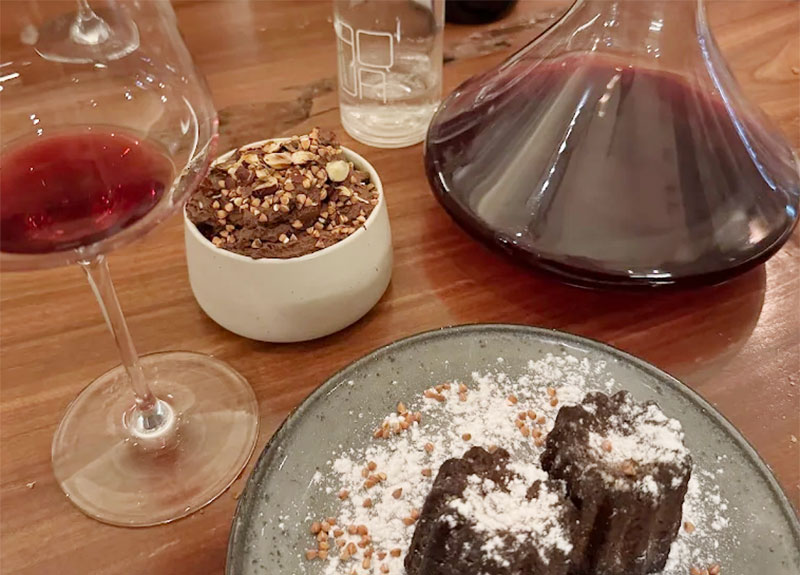 A cozy and inviting scene at Les Amoureuses wine bar in Le Marais, featuring a small wooden table with a glass of red wine, a plate of artisanal cheese and charcuterie, and warm, dim lighting.