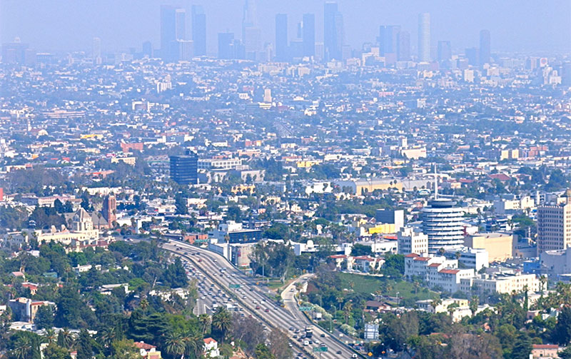 A long shot looking down a major, non-elevated freeway in Los Angeles during rush hour, showing four lanes of heavy, slow-moving traffic with both red and white lights under a hazy sky at dusk.